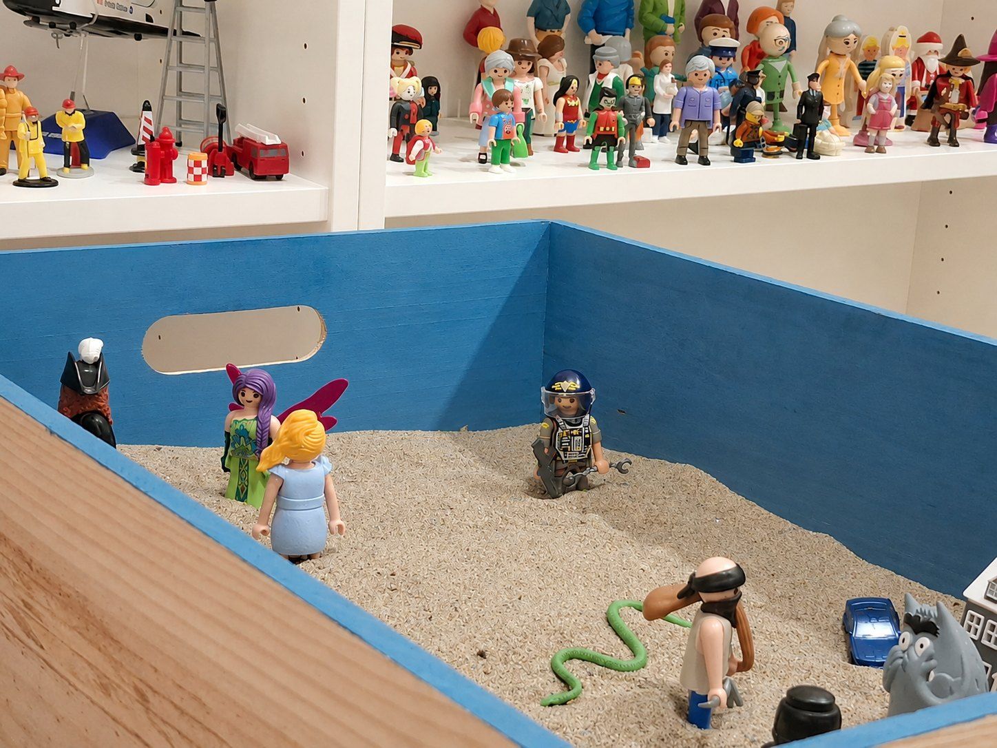 Caja de arena con figuras dispuestas por un niño, y las estanterías de muñecos detrás