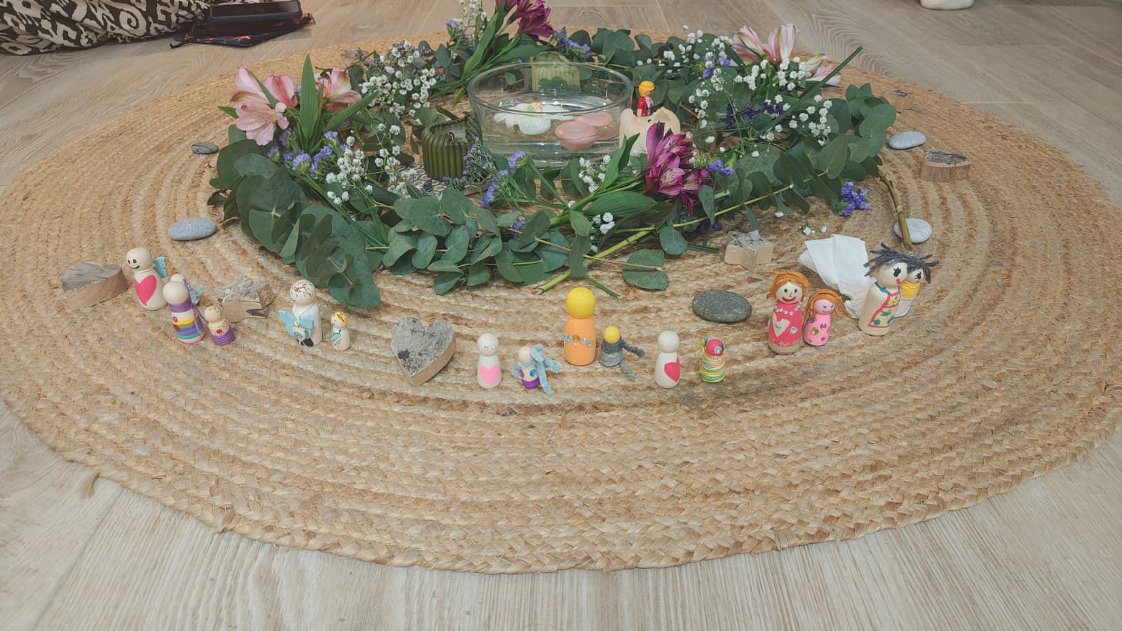 Corona de flores y eucalipto con un cuenco de agua y velas, y figuritas de madera pintadas alrededor, sobre una alfombra de yute
